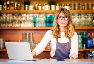 Confident mature small cafe owner businesswoman standing behind the counter and using laptop while working.